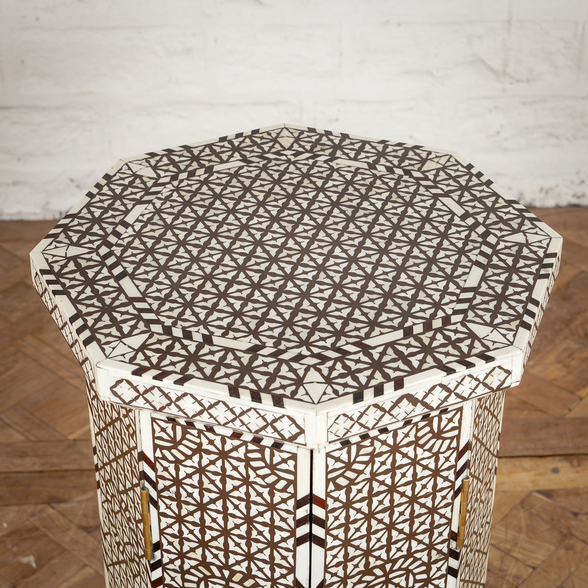 The Karigar Table by John Robshaw is an octagonal wooden table featuring an intricate geometric inlay design in white and brown, handcrafted from premium Indian hardwood and inspired by traditional bone inlay furniture, displayed against a white brick wall and wooden floor.