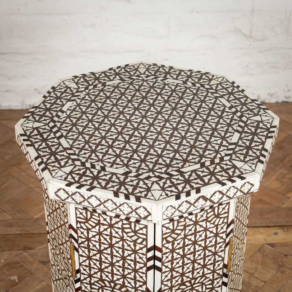 The Karigar Table by John Robshaw is an octagonal wooden table featuring an intricate geometric inlay design in white and brown, handcrafted from premium Indian hardwood and inspired by traditional bone inlay furniture, displayed against a white brick wall and wooden floor.