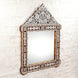 The Ila Bone Inlay Mirror by John Robshaw features a hand-carved frame with intricate floral and geometric mosaic patterns, beautifully mounted on a white textured brick wall. This statement piece adds artistic flair to any space.