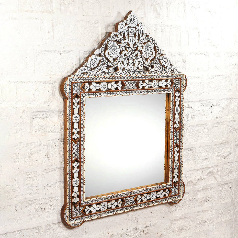 The Ila Bone Inlay Mirror by John Robshaw features a hand-carved frame with intricate floral and geometric mosaic patterns, beautifully mounted on a white textured brick wall. This statement piece adds artistic flair to any space.