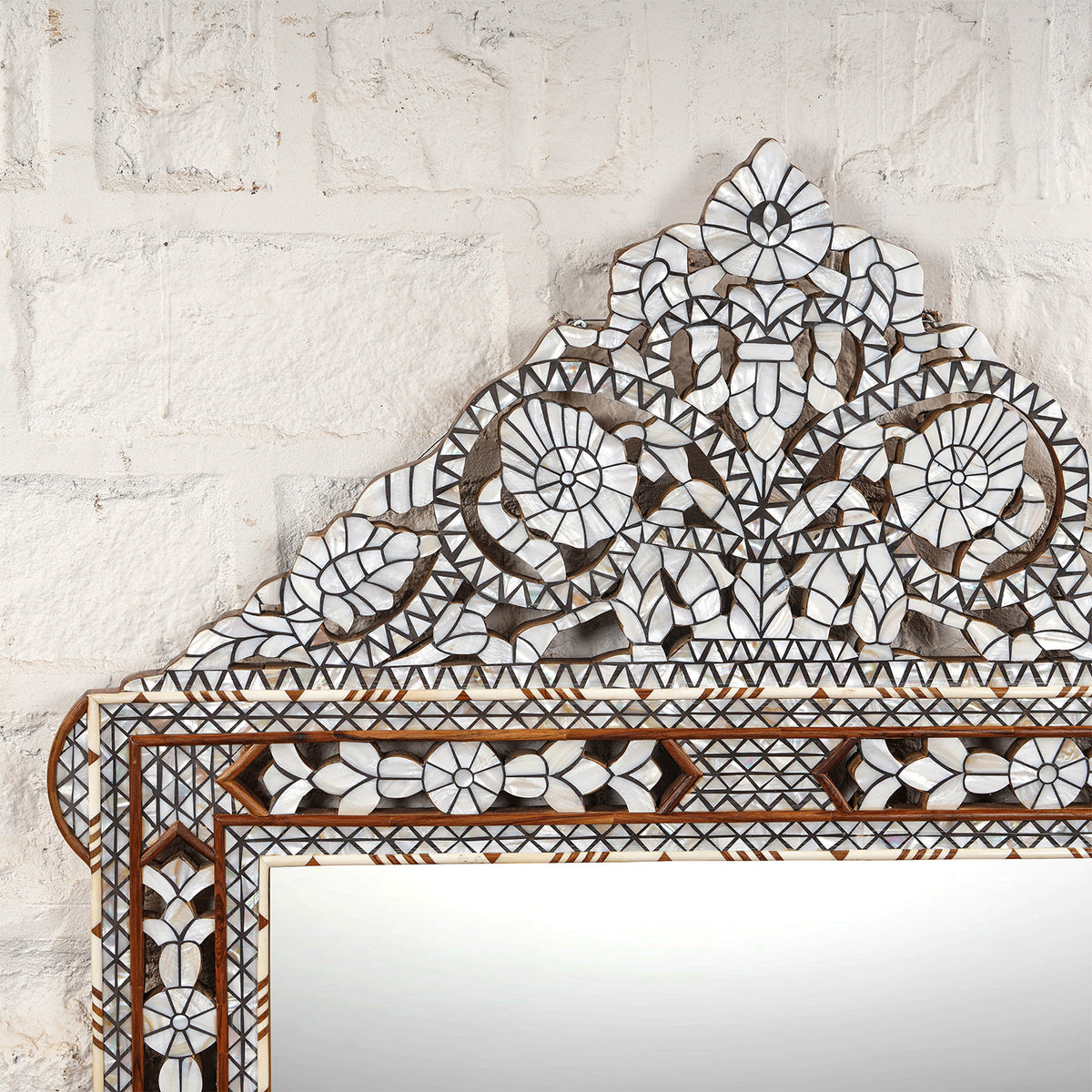 Presenting the Ila Bone Inlay Mirror by John Robshaw—an elegant statement piece featuring intricate geometric and floral inlay work on a hand-carved frame, beautifully set against a white textured brick wall.