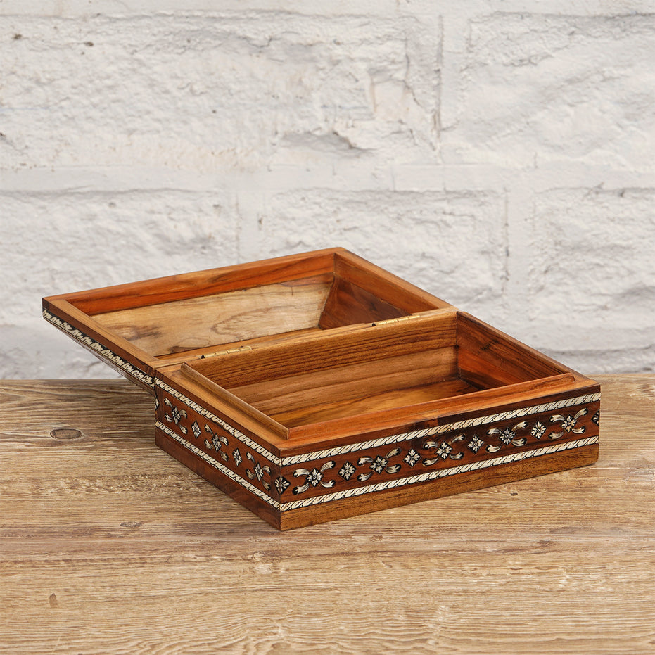 The Sita Bone Inlay Box by John Robshaw is a rectangular wooden box featuring subtle floral patterns and decorative bone inlay. Hinged open to reveal two compartments, it sits atop a teak wood surface against a textured white wall, adding an elegant touch to the display.