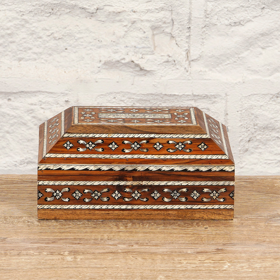 The Sita Bone Inlay Box by John Robshaw features a rectangular teak wood construction with intricate bone inlay patterns in white and brown—some delicately formed into floral motifs—beautifully displayed on a wooden surface against a white brick wall backdrop.