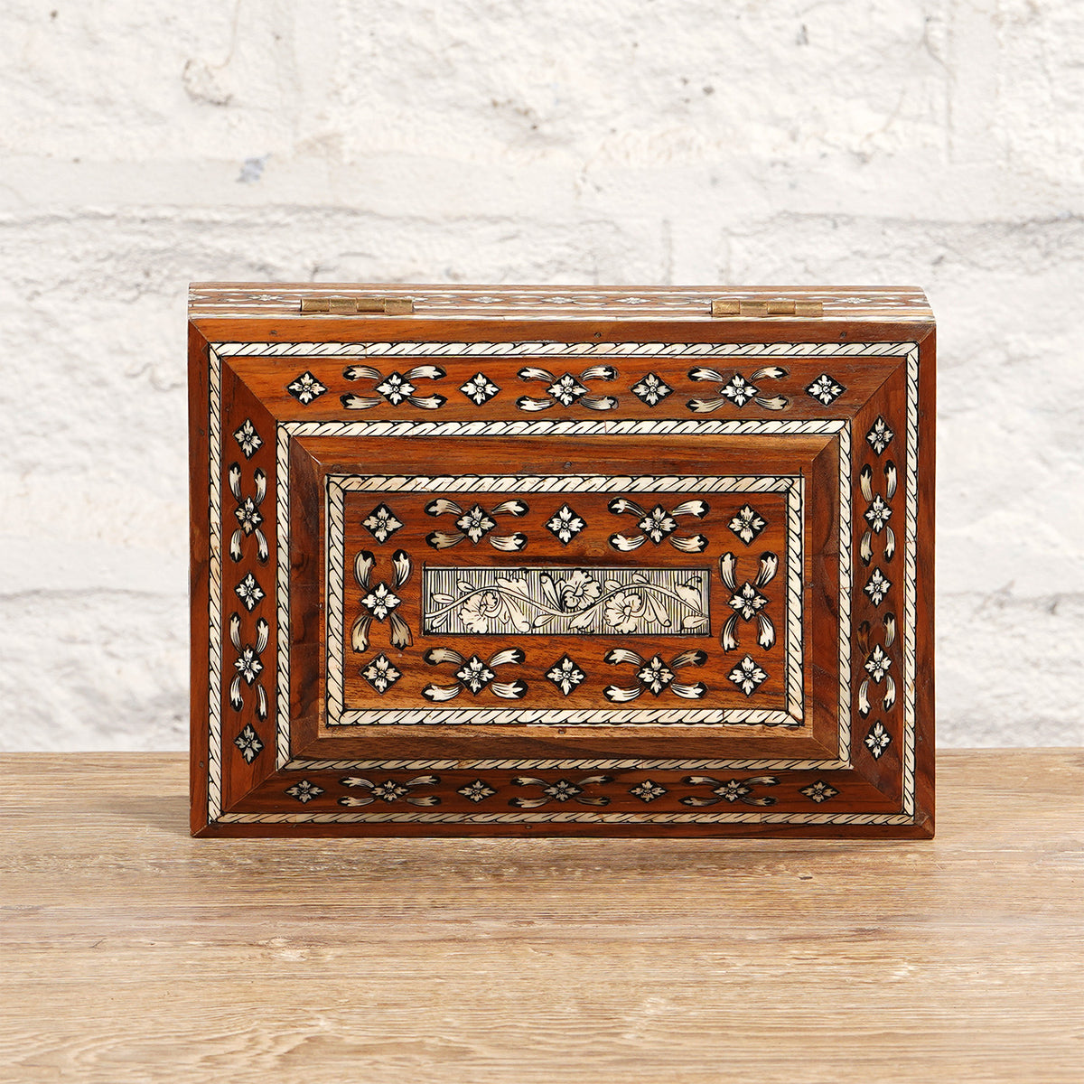 The Sita Bone Inlay Box by John Robshaw, featuring rectangular teak wood construction with intricate bone inlay in floral and geometric patterns, is showcased on a wooden surface against a white brick wall.