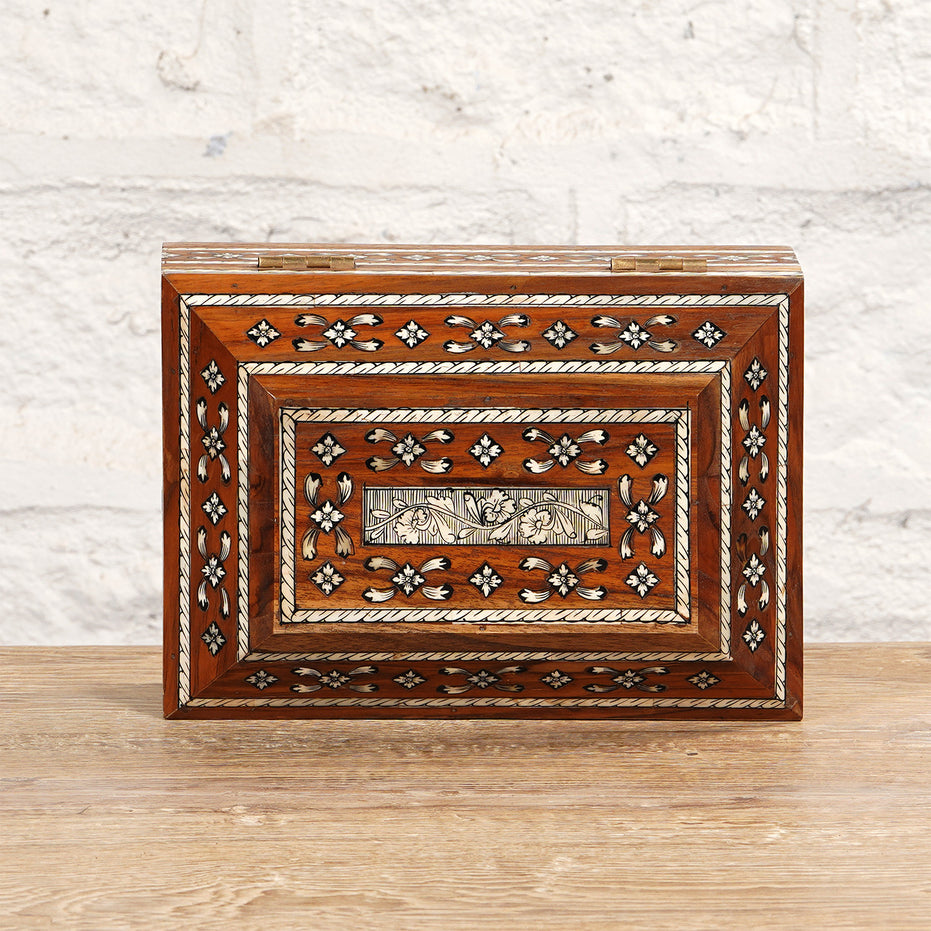 The Sita Bone Inlay Box by John Robshaw, featuring rectangular teak wood construction with intricate bone inlay in floral and geometric patterns, is showcased on a wooden surface against a white brick wall.