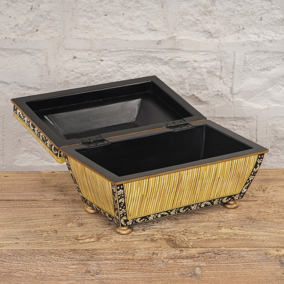 The Suvarna Bone Inlay Box by John Robshaw features a rectangular design with an open hinged lid, adorned with elegant gold and black detailing, and resting on small round feet. Placed on a wooden surface against a light brick wall, it serves as the perfect decorative storage box for your cherished keepsakes.