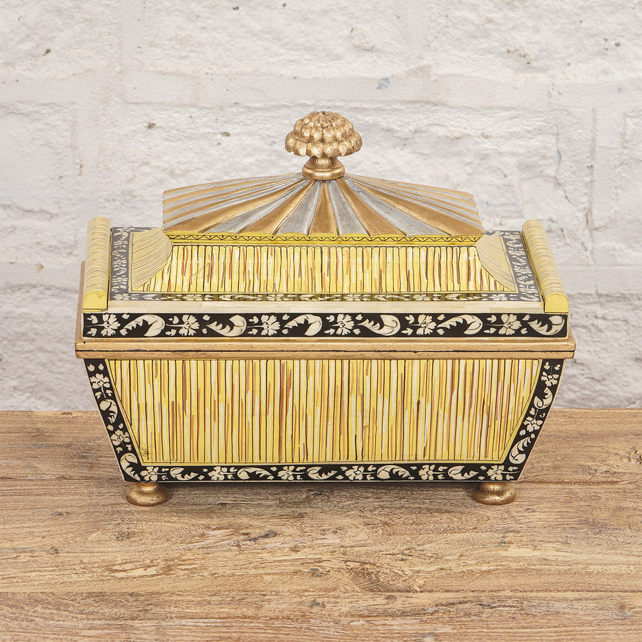 The Suvarna Bone Inlay Box by John Robshaw features a gold and black floral design, ribbed side panels, and a domed lid crowned with a gold finial. Resting on a wooden surface against a white brick wall, this striking piece is reminiscent of an Indian teak box.