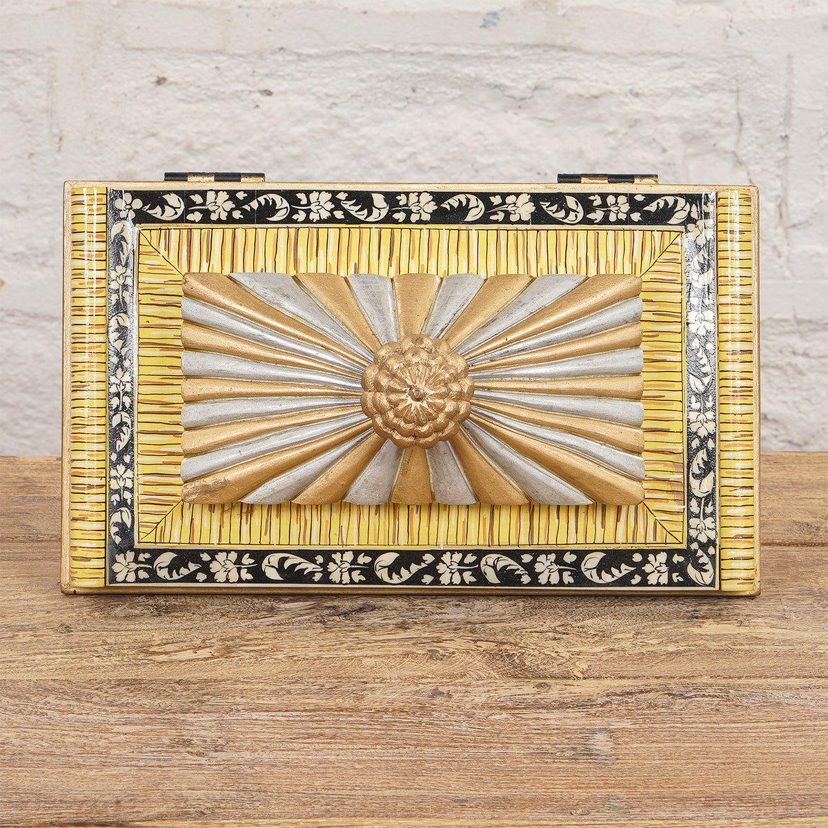 The Suvarna Bone Inlay Box by John Robshaw features a decorative combination of gold, silver, and black accents with a raised floral and sunburst design on the lid, beautifully displayed on a wooden surface against a white brick wall.