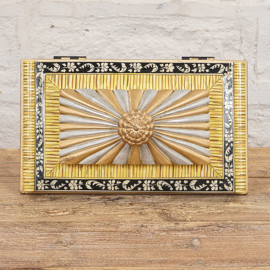 The Suvarna Bone Inlay Box by John Robshaw features a decorative combination of gold, silver, and black accents with a raised floral and sunburst design on the lid, beautifully displayed on a wooden surface against a white brick wall.