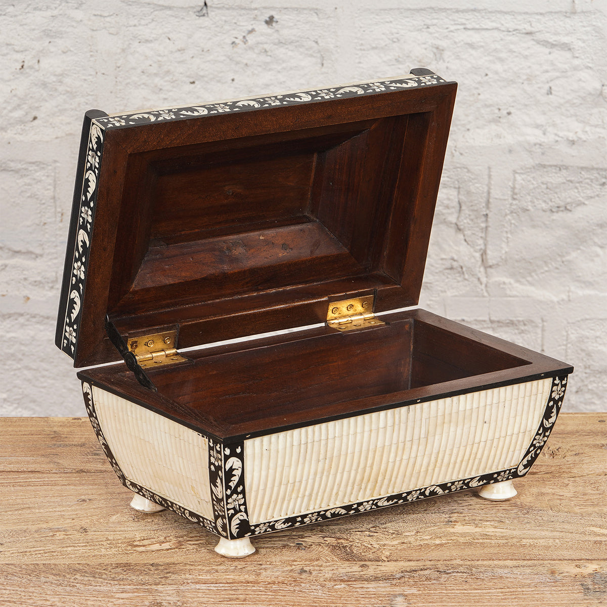 The Lakshmi Bone Inlay Box by John Robshaw, an open decorative teak wood piece adorned with ivory-colored inlays and floral patterns, sits on a wooden surface against a white brick wall, bringing a touch of Indian decor to the space.