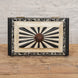 The Lakshmi Bone Inlay Box by John Robshaw features a rectangular shape with a striking black and white radial pattern on the lid, a floral border, and a round central knob. Placed on a wooden surface against a textured white wall, this elegant piece is perfect for adding an exquisite touch of Indian decor to your space.