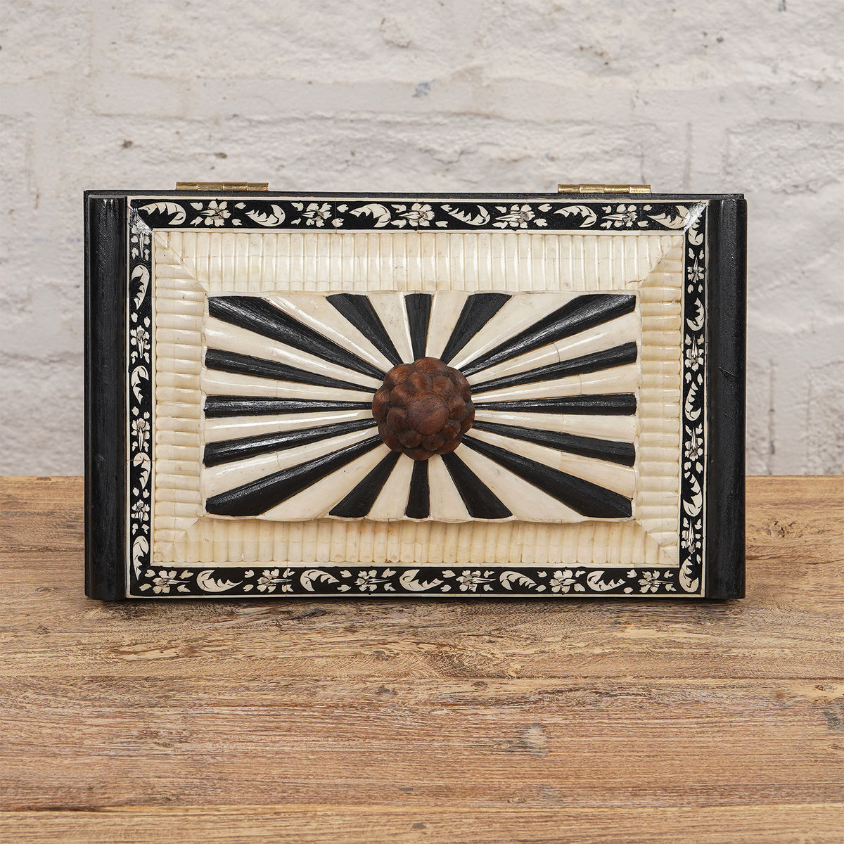 The Lakshmi Bone Inlay Box by John Robshaw features a rectangular shape with a striking black and white radial pattern on the lid, a floral border, and a round central knob. Placed on a wooden surface against a textured white wall, this elegant piece is perfect for adding an exquisite touch of Indian decor to your space.