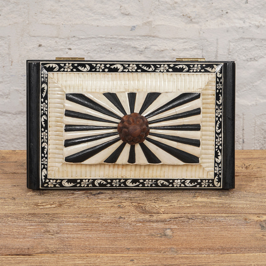 The Lakshmi Bone Inlay Box by John Robshaw features a rectangular shape with a striking black and white radial pattern on the lid, a floral border, and a round central knob. Placed on a wooden surface against a textured white wall, this elegant piece is perfect for adding an exquisite touch of Indian decor to your space.
