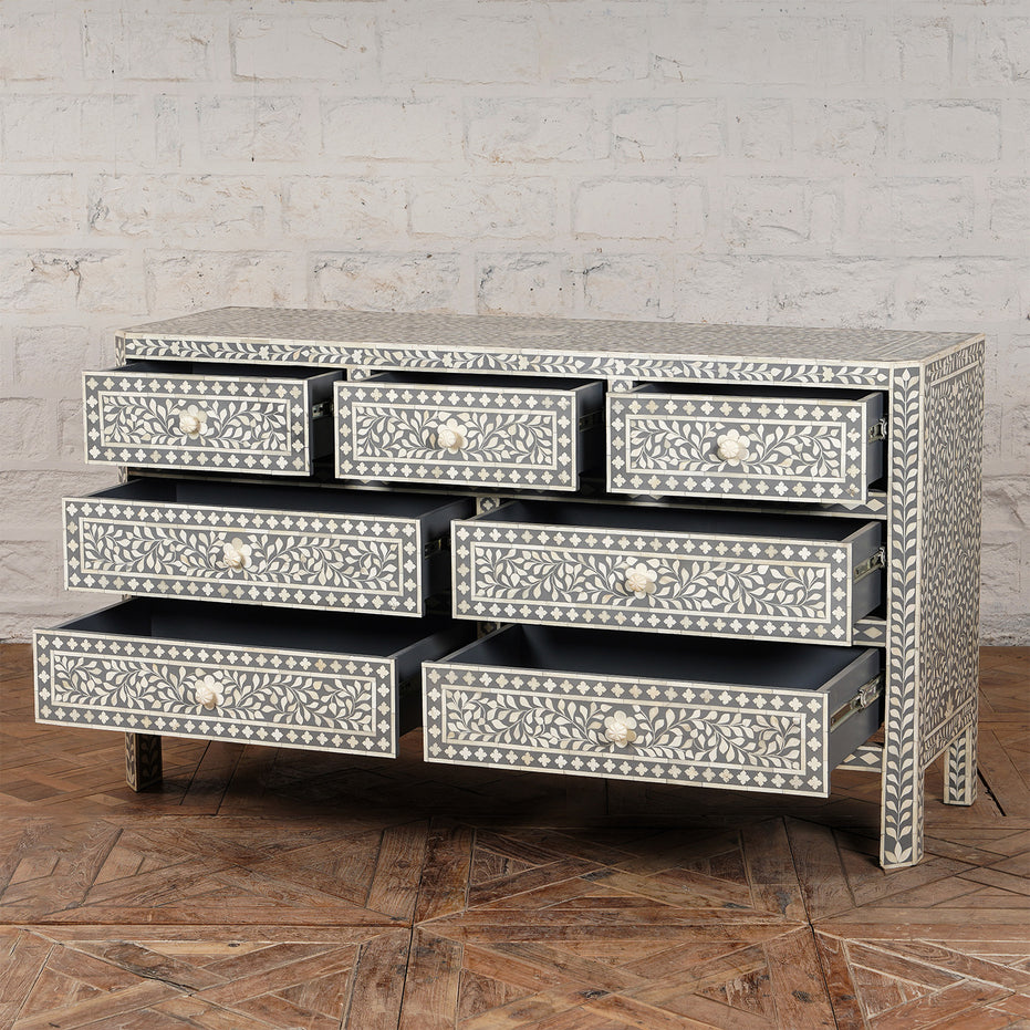 The Vajra Bone Inlay Sideboard by John Robshaw features eight floral-patterned drawers, each partially open, and stands on a wooden floor against a white brick wall. This exquisite piece is both artistic and functional, serving as a true statement in any space.