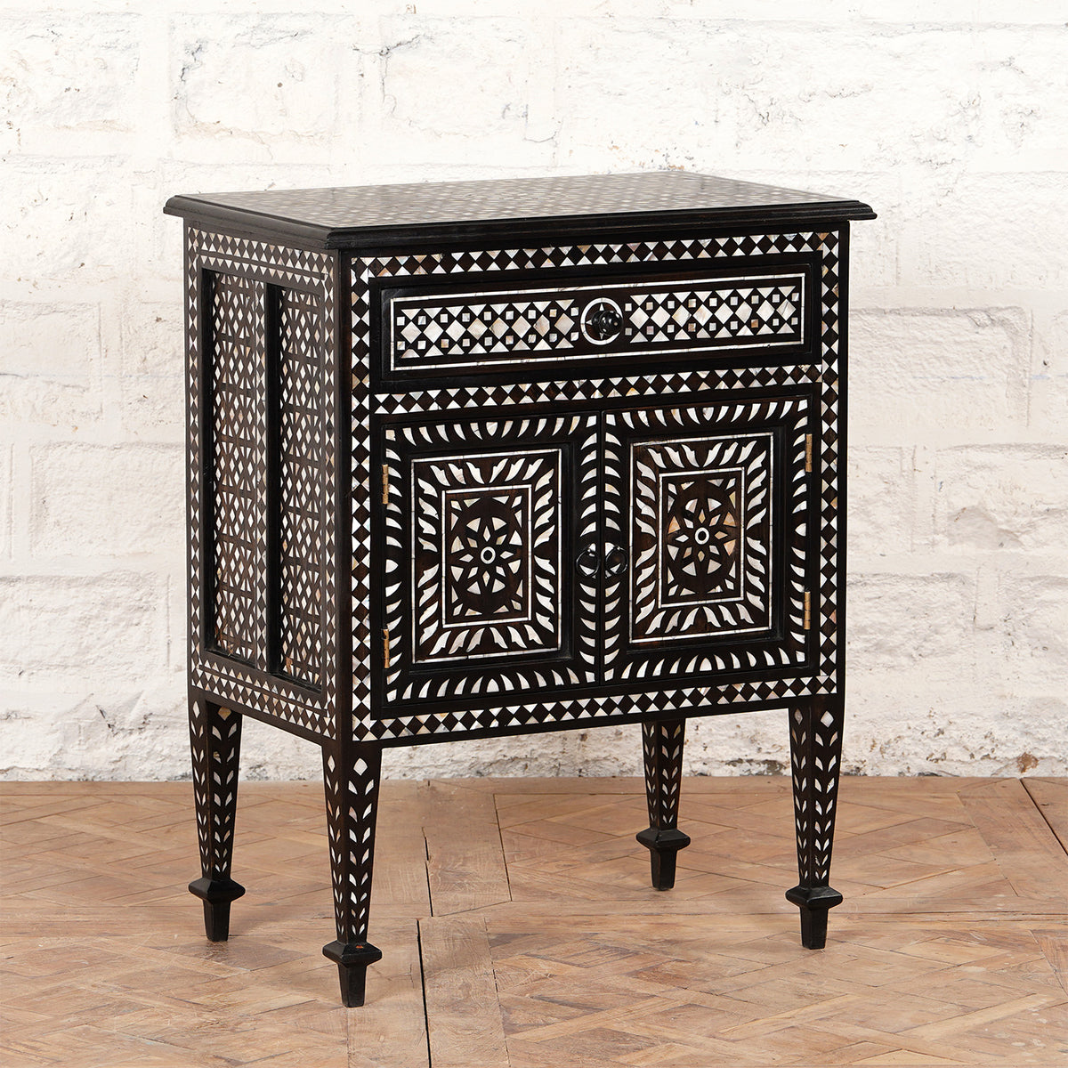 Introducing the Ahan Bone Inlay Cabinet by John Robshaw: a black wooden nightstand handcrafted to perfection, showcasing intricate white geometric inlay patterns. It features one drawer and two cabinet doors and stands elegantly on four tapered legs, pictured here against a white brick wall.