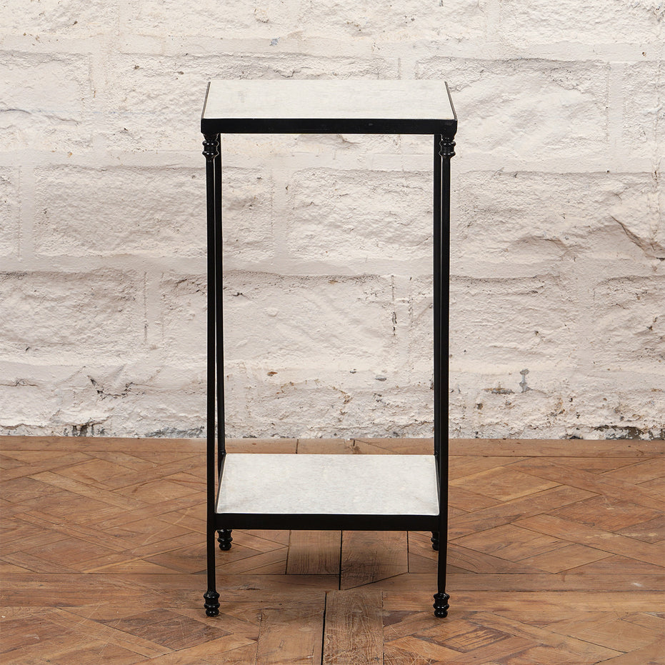 The Veda Side Table by John Robshaw is a small, rectangular two-tier minimalist piece, featuring a black iron frame with light-colored surfaces, shown here standing on a parquet wood floor against a white brick wall.