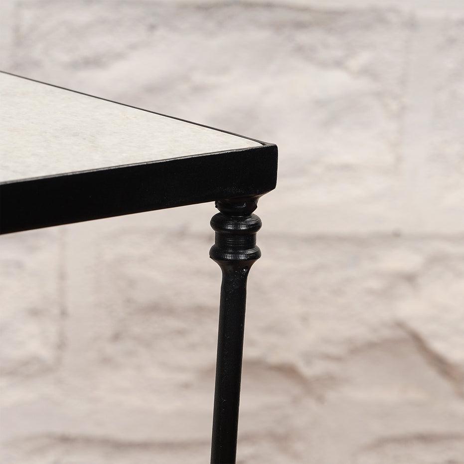 Close-up of a black metal leg with a decorative design on the John Robshaw Veda Side Table, supporting its rectangular tabletop against a light stone wall background, showcasing the minimalist elegance characteristic of this iron side table.