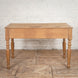 The Smriti Teak Desk by John Robshaw, featuring a rectangular top and four turned legs, stands on a patterned wooden floor against a white brick wall.