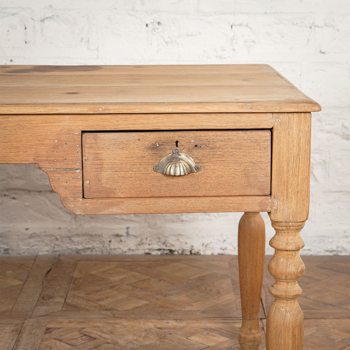 Close-up of the Smriti Teak Desk by John Robshaw, a vintage-inspired writing desk with a single drawer, a distinctive metal shell-shaped handle, turned leg detailing, and charming signs of wear, set against a white brick wall.