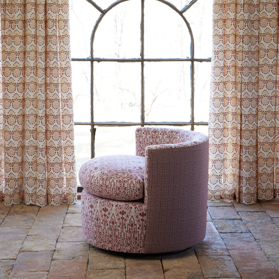 A John Robshaw Round Swivel Chair in Marmar Berry and Aleppo Berry, upholstered in patterned pink performance fabric, sits on a stone floor before a large arched window adorned with matching patterned curtains.