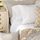 A neatly made bed with crisp John Robshaw Anketi Natural Organic Sheet Set, patterned pillows, a beige throw blanket, and a cream nightstand holding a book and flowers.
