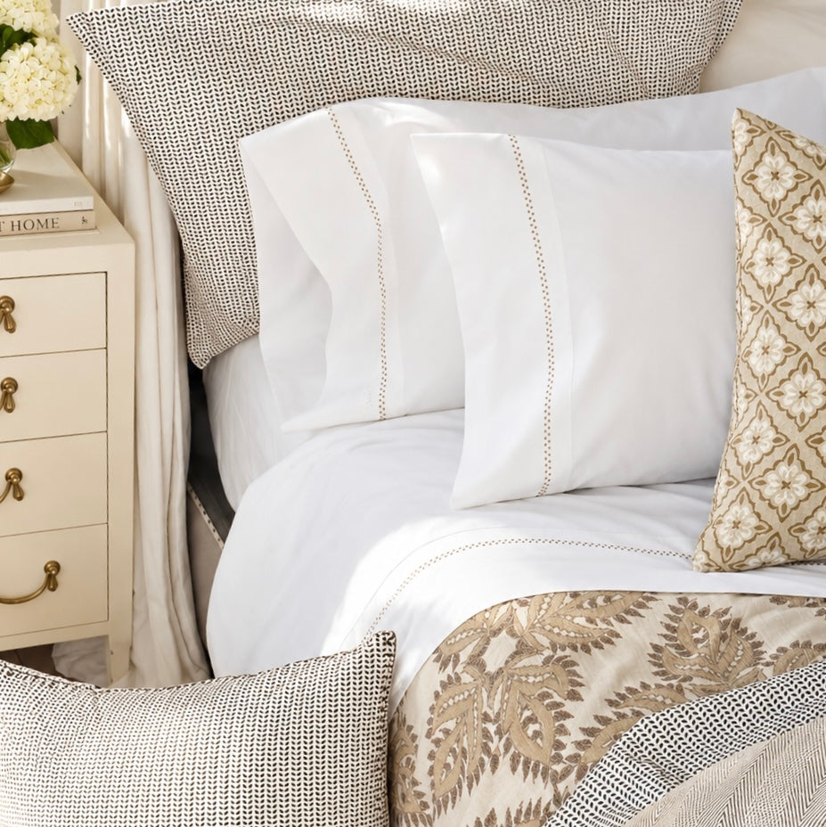 A neatly made bed with crisp John Robshaw Anketi Natural Organic Sheet Set, patterned pillows, a beige throw blanket, and a cream nightstand holding a book and flowers.