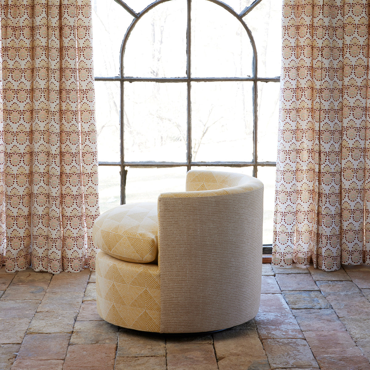 A John Robshaw Round Swivel Chair in Farzu Marigold and Bijal Sand sits on a tile floor in front of a large arched window framed by patterned curtains.