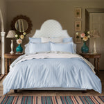 A neatly made bed featuring the John Robshaw Cinde Light Indigo Organic Duvet, flanked by wooden side tables holding vases of flowers and lamps, in a decorated bedroom.