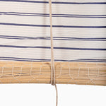 A handmade bamboo Lapis Stripe Chik Blind by John Robshaw Textiles is partially rolled up, secured with a string, against a background of fabric with horizontal blue stripes. - 31884734169134