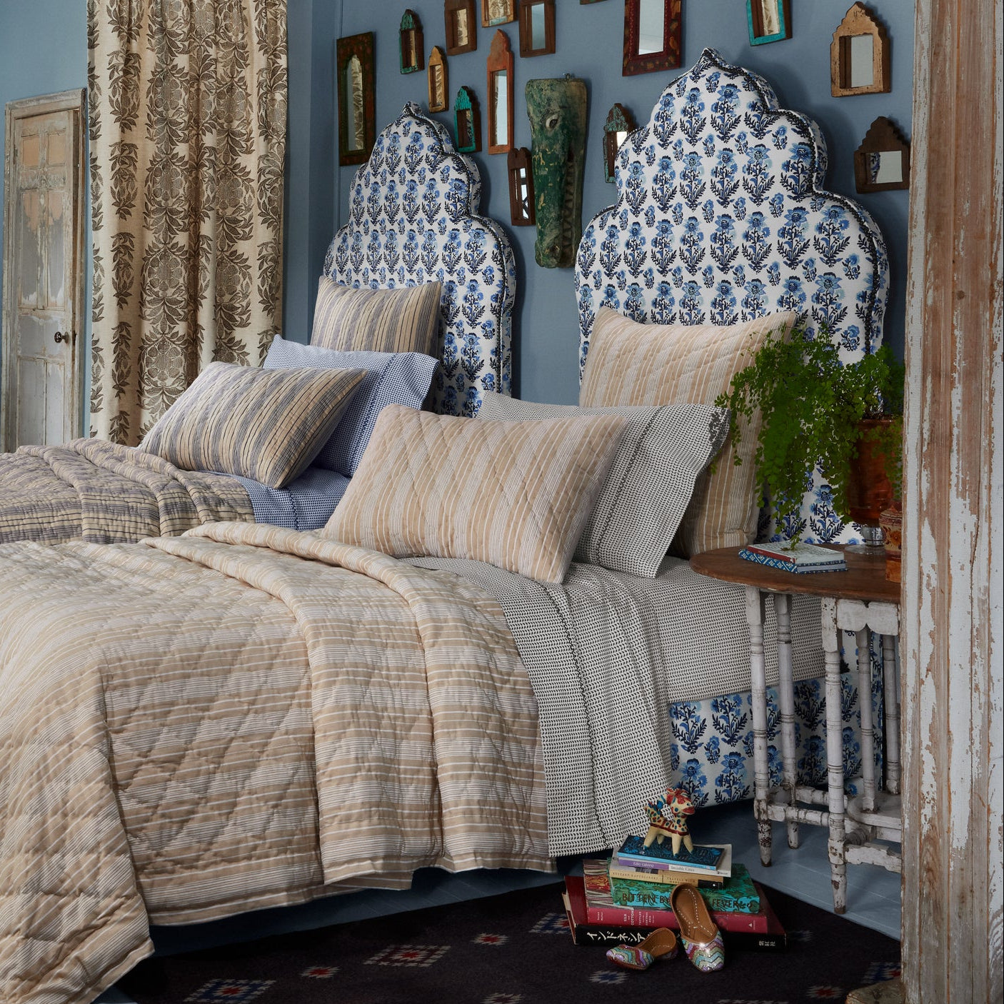 Two beds with patterned blue and white headboards are dressed in John Robshaw’s Cinde Coffee Organic Sheet Set—striped, coffee-print bedding made from 200 thread count organic cotton percale. A bedside table holds books and a plant, while wall-mounted mirrors above reflect the blue wall backdrop.