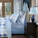 A blue and white bedroom features a patterned quilt, matching pillows, an upholstered bed layered with the Cinde Navy Organic Sheet Set by John Robshaw, a wood nightstand with inlay, and a blue lamp beside large windows with tree views.