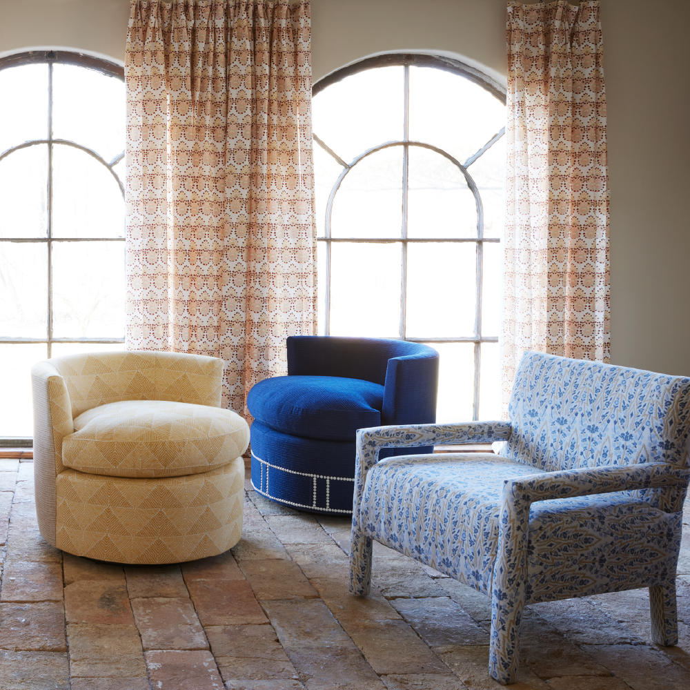 Three John Robshaw Round Swivel Chairs, upholstered in Farzu Marigold and Bijal Sand durable performance fabrics with distinct colors and patterns, are arranged on a tiled floor in front of two arched windows adorned with patterned curtains.