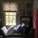 A window nook with a cushioned bench and assorted patterned pillows features Bipin Tangerine Chik Blinds by John Robshaw Textiles. Framed artworks adorn the wall, and trees are visible outside. - 31890988957742