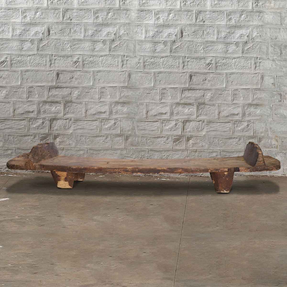 A low, rustic Wooden Naga Coffee Table 8 by John Robshaw—with short legs and a weathered surface evocative of antique tribal craftsmanship—is placed on a concrete floor against a light gray brick wall.