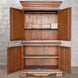 An Anglo Indian Teak Inlaid Cabinet by John Robshaw, featuring ornate trim, two shelves, and a small drawer, stands open against a gray brick wall—an exquisite piece evocative of classic bone inlay furniture.