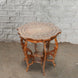 The Round Wooden Inlay Table 10 by John Robshaw features an ornate bone inlay design, decorative legs, and a lower shelf, beautifully crafted from Indian hardwood. Shown here on a concrete floor against a light gray brick wall.