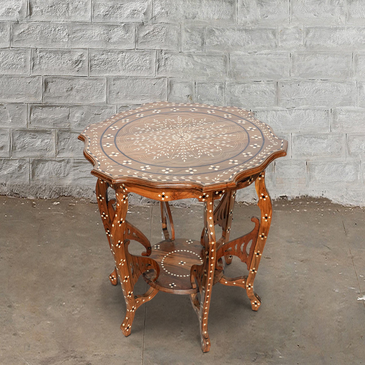 The Round Wooden Inlay Table 10 by John Robshaw features an ornate bone inlay design, decorative legs, and a lower shelf, beautifully crafted from Indian hardwood. Shown here on a concrete floor against a light gray brick wall.