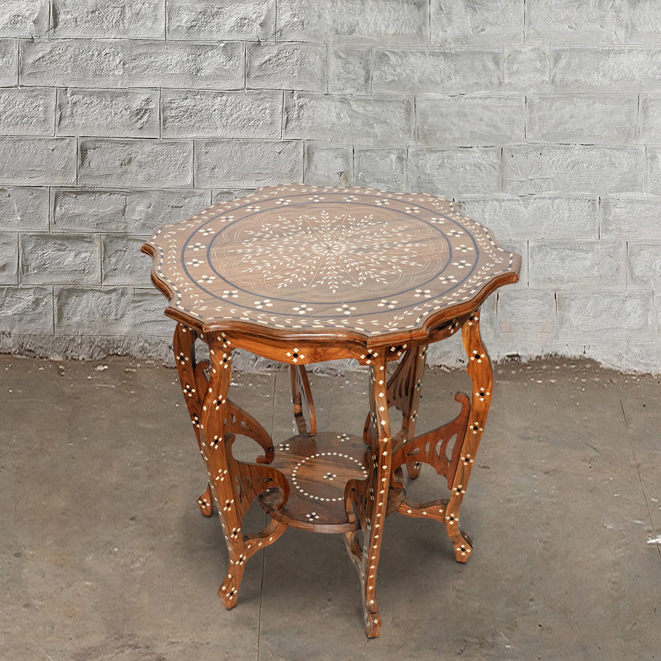 The Round Wooden Inlay Table 10 by John Robshaw features an ornate bone inlay design, decorative legs, and a lower shelf, beautifully crafted from Indian hardwood. Shown here on a concrete floor against a light gray brick wall.