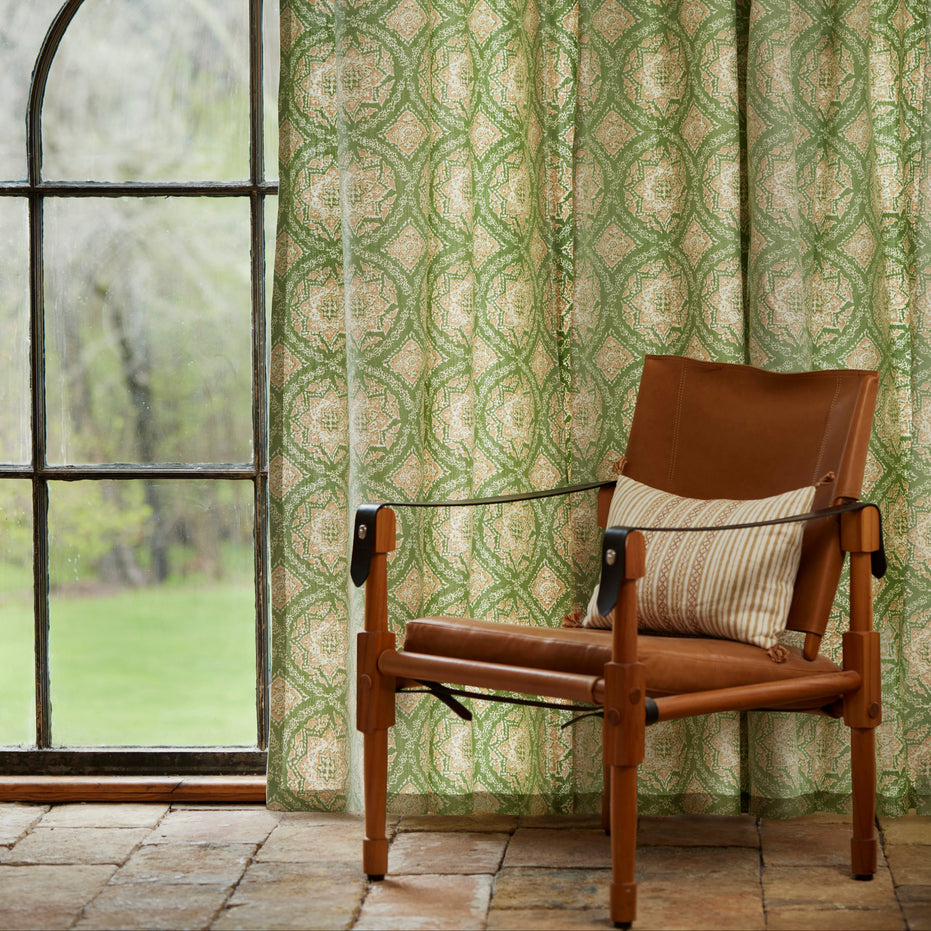 A brown leather chair with a striped pillow sits on a tile floor by a large window, adorned with John Robshaw’s Maansi Sage curtains featuring a classic medallion print.
