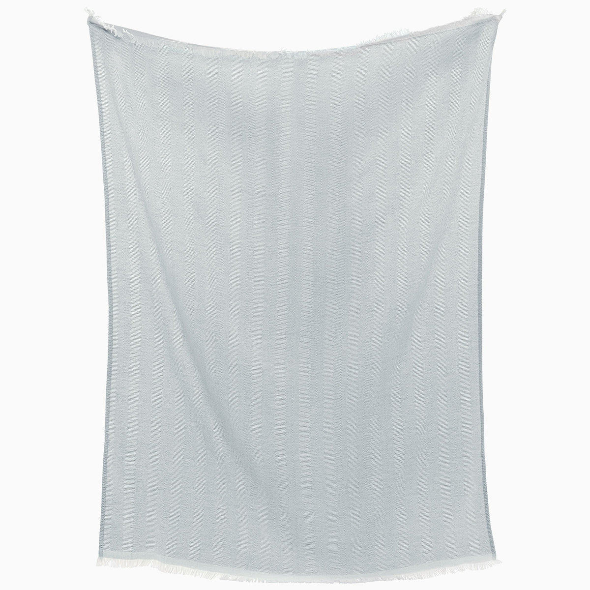 The Lavanya Mist Throw by John Robshaw, a light blue rectangular fabric with fringed edges and subtle herringbone weave, hangs against a white background.