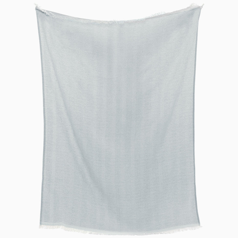 The Lavanya Mist Throw by John Robshaw, a light blue rectangular fabric with fringed edges and subtle herringbone weave, hangs against a white background.