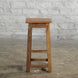 The Ambu Teak Stool by John Robshaw, featuring a solid teak construction and square seat, stands on a concrete floor against a white brick wall, capturing the timeless appeal of vintage furniture.