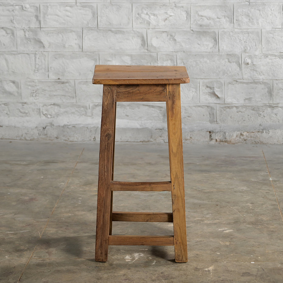 The Ambu Teak Stool by John Robshaw, featuring a solid teak construction and square seat, stands on a concrete floor against a white brick wall, capturing the timeless appeal of vintage furniture.