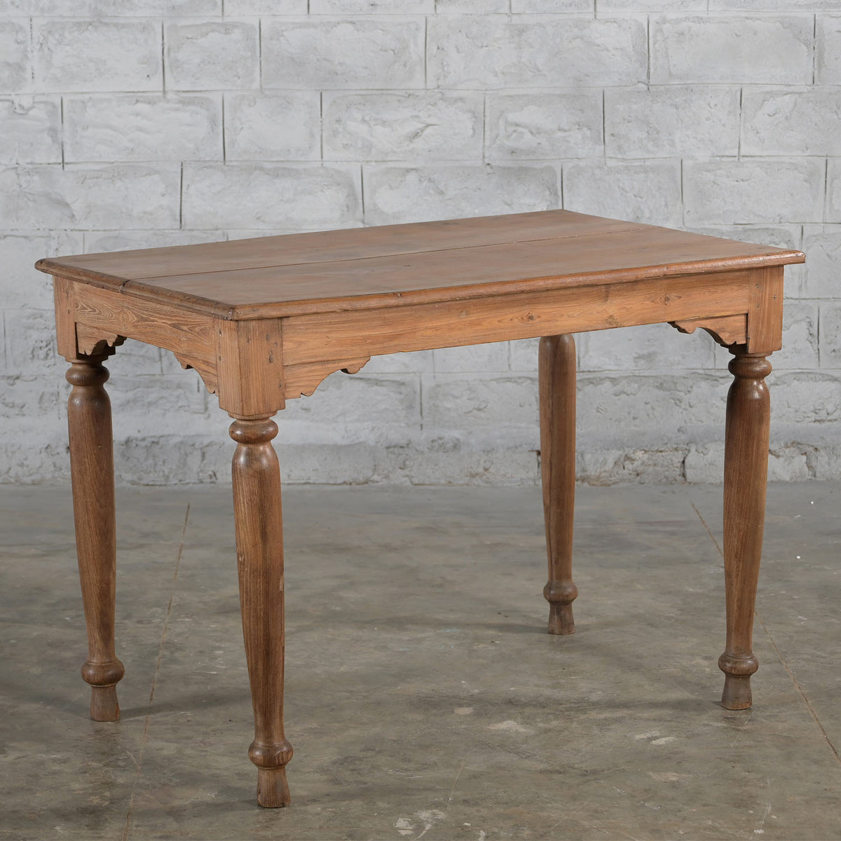 The Hanita Teak Table by John Robshaw, featuring a handcrafted rectangular design with four turned legs, stands on a concrete floor against a light gray brick wall background, evoking the charm of vintage furniture.