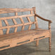A John Robshaw Oorja Teak Bench, with angled back slats and a weathered finish showing visible wear, sits on a concrete floor against a gray brick wall.