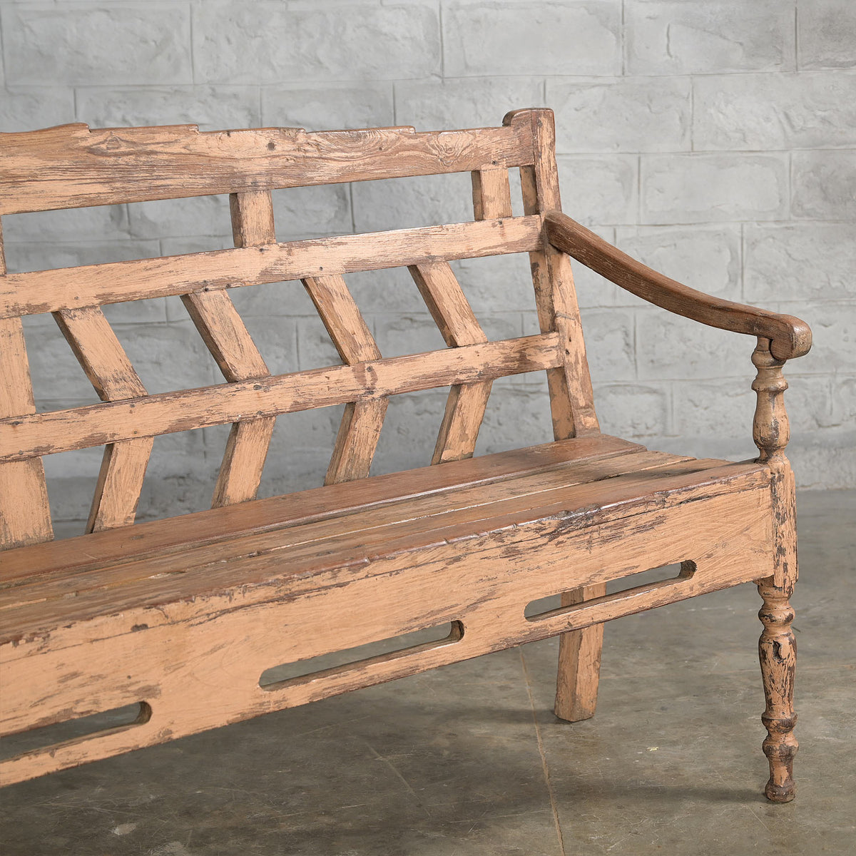 A John Robshaw Oorja Teak Bench, with angled back slats and a weathered finish showing visible wear, sits on a concrete floor against a gray brick wall.
