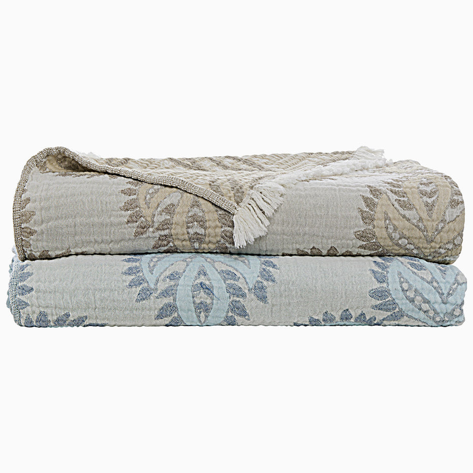 Two folded patterned blankets, one brown and one blue, stacked on top of each other. The top blanket, the Dasati Sand Throw from John Robshaw, is crafted from stonewashed cotton and also features fringe detailing.