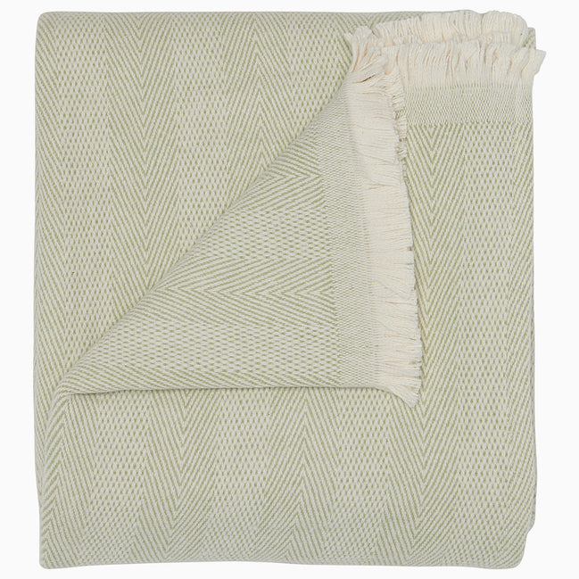 The Lavanya Sage Throw by John Robshaw is a folded, light cotton blanket featuring a subtle herringbone pattern and one fringed edge.