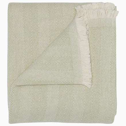 The Lavanya Sage Throw by John Robshaw is a folded, light cotton blanket featuring a subtle herringbone pattern and one fringed edge.