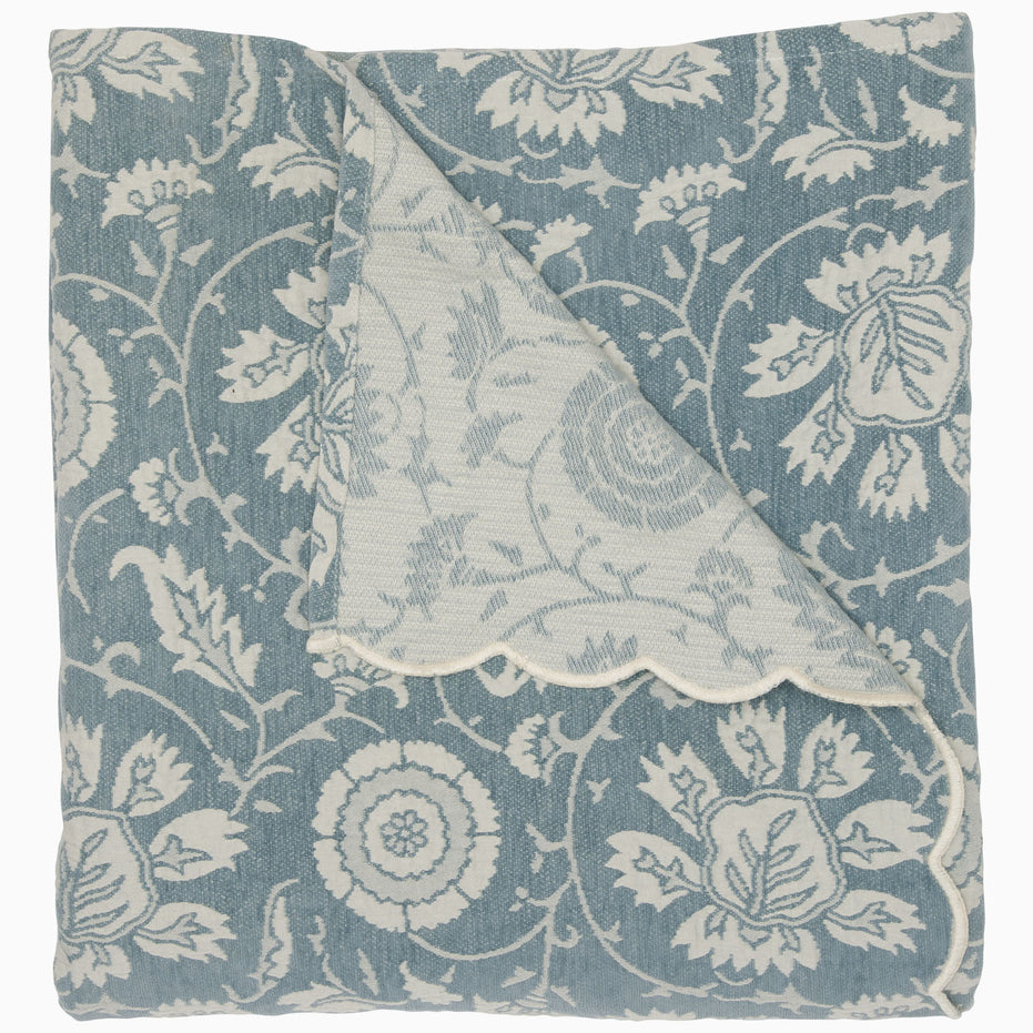 A partially unfolded Kimari Mist Coverlet by John Robshaw, showcasing its delicate woven floral patterns and scalloped edges in blue and white.